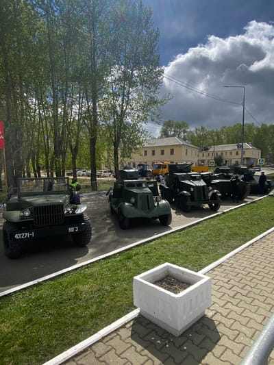 A collection of vintage military vehicles are parked in a line along a street in Kirovgrad, Russia. The vehicles include trucks and armored cars, all in a dark green color. Some people are visible in and around the vehicles. The setting is an urban environment, with a row of trees on one side of the street, and a building on the other. The sky is partly cloudy. A flower bed is visible in the foreground on the right. The street is paved with interlocking concrete pavers.