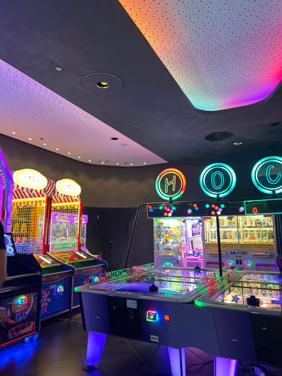 This image captures a vibrant, modern indoor arcade or entertainment center in Agios Dimitrios, Greece, characterized by its colorful artificial lighting and diverse game machines. The scene suggests an evening or nighttime setting due to the reliance on artificial illumination.

In the foreground and extending into the midground, several arcade games are visible. These include various prize redemption machines, such as claw games with glass casings displaying prizes and other coin-operated or ticket-redemption games. The machines are brightly lit with internal and external neon elements, showcasing glowing controls and game elements. Text "Hi-Tech" is visible in a neon-style font on at least two game machines. Other visible text on the side of machines includes "RE 1012" and "RE 1047".

The architectural design of the venue is notable. A large, curved ceiling or wall section on the left features a textured, perforated surface illuminated by a gradient of rainbow colors, transitioning from soft pinks and purples to blues, greens, yellows, and oranges. This creates a striking visual effect. Adjacent to this, a darker, smooth wall or ceiling area has circular cutouts, some housing lights or vents.

Prominently displayed on a dark background in the upper right portion of the image are three large, circular neon signs, glowing brightly with different colors. They depict the stylized letters "C", "Q", and "H" from top to bottom.

The overall atmosphere is one of dynamic leisure and excitement, typical of an entertainment complex. No individuals are present in the frame, allowing the focus to remain on the striking visual elements and modern aesthetic of the arcade.