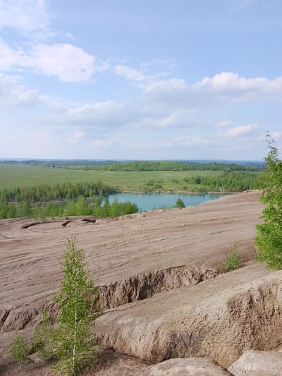 The image depicts a landscape in Uzlovaya, Russia. In the foreground, there's a brown, terraced, eroded terrain that resembles a quarry or mining site. Sparse vegetation, including small birch trees, dots the landscape. In the midground, a body of water, likely a lake or pond, displays a light blue color. Behind the water, a line of green trees and forest extends into the distance. A flat expanse of green fields lies beyond the trees. The sky above is light blue with scattered white clouds. There are no people visible in the image.