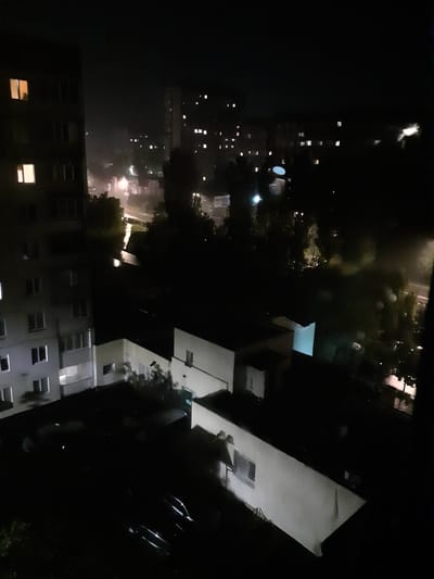 This is a night-time, aerial view of a dimly lit residential area in Obukhiv, Ukraine. The scene is dominated by the dark shapes of apartment buildings and trees, with scattered lights from windows and street lamps illuminating the night. In the foreground on the left is a tall apartment building with several illuminated windows. To the right of this building, there are lower structures, also with some lights visible, and a street or pathway that appears wet and reflective, suggesting recent rain or a puddle. Further into the distance, more apartment buildings with illuminated windows are visible, set against the dark sky. The overall impression is one of a quiet, overcast night in a city neighborhood.