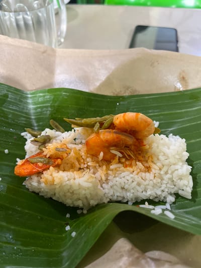 A plate of rice with shrimp and green beans is served on a banana leaf, which is placed on a brown paper wrapper. The food is topped with a reddish sauce. In the background, a glass of water and a smartphone are visible. The scene appears to be in a restaurant or eatery in Manyang Cut, Indonesia.