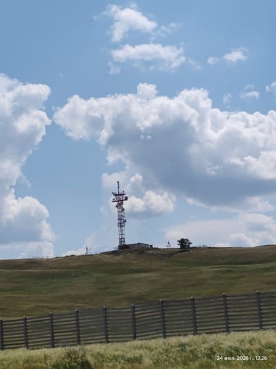 The image shows a telecommunications tower on a grassy hill under a partly cloudy sky. In the foreground, a wooden fence runs horizontally across the frame. The scene is located in Keneсары, Kazakhstan, as indicated by the date stamp "24 июл. 2026 г., 13:26" which translates to "July 24, 2026, 13:26". The tower is a lattice structure with multiple antennas and platforms. There are a few small buildings and a single tree visible on the hill near the tower. The grass on the hill is mostly green and brown, suggesting a summer or early autumn season. The sky is blue with scattered white cumulus clouds.