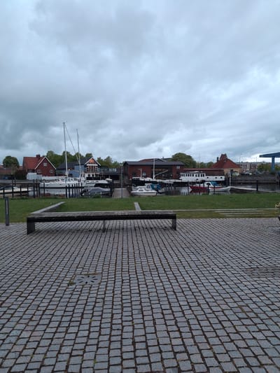 The image depicts a waterfront scene in Køge, Denmark. The foreground is dominated by a paved area constructed of grey cobblestones. A dark metal bench is positioned parallel to the waterfront. Beyond the bench, a strip of green grass separates the pavement from the water. Various boats are moored along the waterfront, including sailboats with tall masts. Buildings with red brick and dark-colored roofs line the waterfront. The sky is overcast with grey clouds.