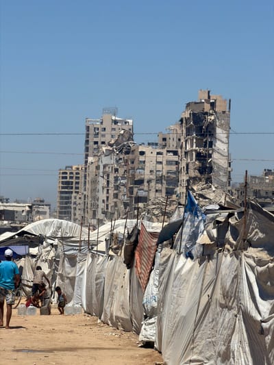 In the city of غزة, Unknown, a scene unfolds under a clear blue sky. In the foreground, a man in a blue t-shirt and patterned shorts walks away from the camera, his back to the viewer. Beside him, a bicycle stands upright. Further into the frame, a group of children are gathered. One child, wearing a patterned top, appears to be interacting with a bicycle. Another child, in a grey tank top and dark shorts, is crouched down. Nearby, a few large, clear plastic water containers are visible, some of them partially filled. The children are surrounded by makeshift tents or shelters constructed from white tarps and fabric, creating a sense of a temporary settlement.

In the background, the stark reality of destruction is evident. Several multi-story buildings stand in ruins, their facades heavily damaged, with gaping holes and debris scattered around them. The architecture suggests apartment buildings or similar structures, now reduced to skeletal remains. The overall environment is dusty and barren, with a dirt path in the foreground. The presence of flags, including one predominantly blue and white, adds a touch of color to the otherwise somber landscape. The contrast between the resilient makeshift shelters and the devastated buildings highlights a scene of displacement and survival amidst destruction.