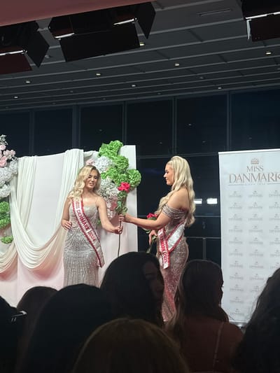 The image captures a scene from a Miss Denmark event. Two women, both blondes, are wearing sequined gowns and sashes labeled "Miss Denmark" with different years (2024 and 2025). The woman wearing the Miss Denmark 2025 sash hands a rose to the woman wearing the Miss Denmark 2024 sash. They are positioned near a white backdrop decorated with floral arrangements. Behind them is a banner with the "Miss Danmark" logo. The background is a modern-looking interior, possibly a hall or auditorium, with a high ceiling and gray vertical lines. The photograph appears to have been taken from an audience perspective, with the heads of other audience members visible in the foreground. Skovshoved, Denmark is written in the prompt so I am adding that location data to the description.