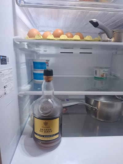 The image shows the inside of a refrigerator. On the left side, there is an egg carton with brown eggs. Adjacent to the eggs, there's a container of what appears to be cheese or yogurt. A stainless-steel pot with a handle is placed near the container. Lying horizontally on the lower shelf is a bottle of "Vega Cadur Brandy de Jerez Gran Reserva" with a cork stopper. The location is implied to be in Xirivella, Spain based on the presence of the Spanish brandy. Additional products in the refrigerator include what appears to be a container of burrata cheese. A "SmartThings" sticker is attached to the interior wall of the fridge.