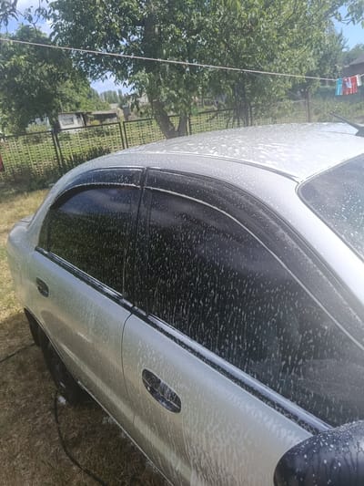 A light-colored sedan, likely being washed, is parked on dry grass outdoors. The vehicle is covered in soap suds. The background reveals a wooden fence, trees with green foliage, and a glimpse of buildings. In the upper right corner, a clothesline with several clothespins is visible. The scene is set on a bright, sunny day. The image is taken from a side-rear angle of the car. The specific location is Pishchane, Ukraine.