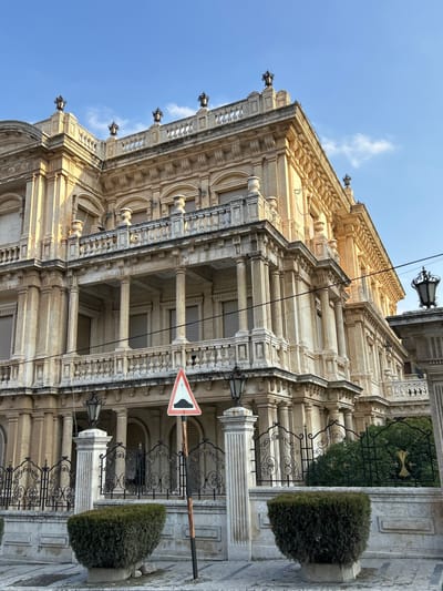 The image displays a grand, ornate building captured from a low-angle perspective under a clear blue sky with a few wispy clouds. The multi-story structure is made of light-colored stone, possibly beige or cream, and features elaborate classical architecture. Prominent details include numerous columns, decorative balustrades, intricate cornices, and balconies. Several ornate street lamps hang from the building's facade, adding to its historical aesthetic.
In the foreground, dark, decorative wrought-iron railings enclose sections of what appears to be a courtyard or sidewalk area, alongside neatly trimmed evergreen bushes planted in large pots. A triangular road sign with a red border, white background, and a black symbol (indicating a "Give Way" or "Yield" instruction) is visible on a pole near the center of the lower half of the image. Utility wires crisscross the sky, partially obscuring the building's upper sections.
The scene is well-lit by natural sunlight, suggesting it is daytime, likely mid-morning or afternoon. The architecture conveys a sense of historical significance and grandeur. The location is identified as Aleppo, Syria. There is no visible human activity, and no decipherable text other than the symbol on the road sign.