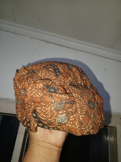 In Na Sang, Laos, a human hand holds a crumpled piece of fabric with a distinct pattern. The fabric, likely a garment or headwear, exhibits a brown and orange base with intricate, swirling, and dotted motifs in lighter beige and darker gray/black, resembling traditional Batik design. The hand, showing a light to medium skin tone, supports the fabric from below, with the palm and part of the wrist visible. The background consists of a plain, light-colored wall (presumably white or off-white) with a horizontal decorative molding or trim running across the upper portion. Below the fabric, to the left and right, dark, indistinct areas are visible, possibly a window or a dark opening, framed by a lighter border. An artificial light source illuminates the scene, casting a prominent shadow of the fabric onto the wall behind it.