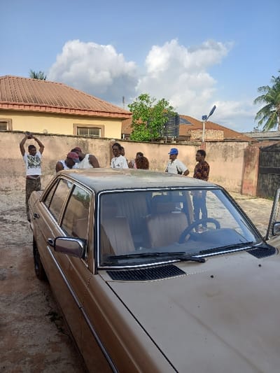The image shows a brown sedan parked in front of a two-story building. There are six people standing around the vehicle. One person is holding a blue umbrella and another is holding a camera. The house is in the background with a white roof and a brown wall. There are trees and a water tank behind the building. The image was taken in Ita Oniyan, Nigeria, located at the coordinates (latitude: 7.286, longitude: 5.16).