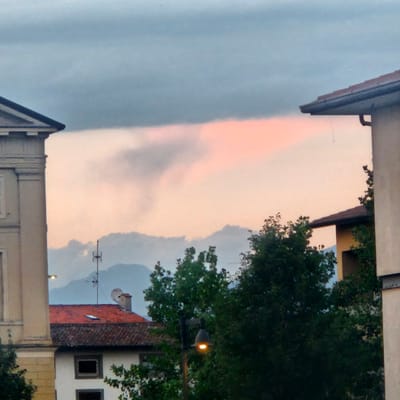 This image, taken in Calcinate, Italy, presents a view of a cloudy sky with a soft pink hue, likely from a sunset or sunrise, illuminating the clouds and the distant mountain range. In the foreground, parts of buildings are visible. On the left, a beige building with architectural detailing and a sloped roof edge is partially in frame. To the right, another building with a lighter colored facade and a dark roof is seen. A dense canopy of green trees dominates the midground. A streetlamp with its light on is positioned in front of the trees. A red-tiled roof of a lower building with a white facade and dark window frames is visible beneath the trees and behind the streetlamp, and this building appears to be part of the urban landscape. Various antennas and satellite dishes are perched on the roof of this building. The overall impression is a serene, dusk or dawn scene in a European town, with a mix of architecture and natural elements.