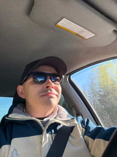The image is an upward-angled shot taken from inside a car, presumably in motion or at a standstill. A person is visible, wearing a dark baseball cap and sunglasses, with a jacket in shades of blue and white. The car features a sunroof and a sun visor with a visible warning label. Outside, the sky is visible through the sunroof, along with trees. Given the language of the warning label, this is unlikely to be in Russia, and is thus not located in Severnyy, Russia. The setting seems to be during daylight hours with bright sunlight.