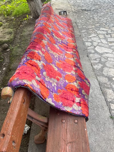 A long, wooden bench is covered with a colorful, floral-patterned blanket in shades of purple, red, and orange. The bench appears to be situated outdoors, with a patch of green grass and a tree trunk visible on the left side of the frame. To the right of the bench is a paved pathway made of irregular grey stones. A small, dark metallic object, possibly a thermos or a container, rests on the far end of the bench. The scene is set in the city of Tashlak, Kyrgyzstan.