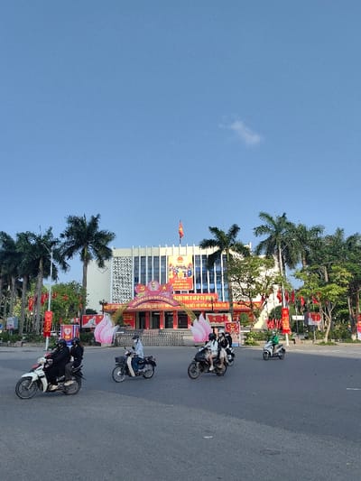 The image depicts an urban street scene in Thuận Hóa Ward, Vietnam, in front of a prominent public building under a clear blue sky with a single small cloud. The foreground features a wide asphalt road where six motorbikes are in motion. Nine individuals are visible on these motorbikes, with most wearing helmets; some also wear face masks.
The central structure is a multi-story building with a light-colored facade, horizontal architectural elements, and numerous windows. A Vietnamese flag with a yellow star on a red field flies from its roof. The building's entrance is heavily decorated for a political celebration. A large archway, featuring a yellow star and hammer-and-sickle emblem, spans the entrance, bearing Vietnamese text translated to "Celebrating the 92 glorious years of the Communist Party of Vietnam (3/2/1930 - 3/2/2022)" and "Welcome to the National Assembly Congress." Pink lotus flower decorations flank the archway. Red banners with yellow Vietnamese text are displayed on the building's facade and along the street, alongside posters featuring Ho Chi Minh.
Tall palm trees line the street on both sides of the building, with additional green foliage to the right. A dark metal fence separates the building complex from the street. One motorbike on the left carries two individuals in dark attire; the rider wears a white helmet, the passenger a dark helmet. In the center, a rider in a white shirt and dark helmet operates a dark motorbike. Another dark motorbike to the right carries two individuals in light clothing and dark helmets. Further right, a solo rider in a bright green top, possibly a delivery service uniform, is on a white scooter, followed by another solo rider on a dark motorbike. All motorbikes are on the paved road, moving in various directions.