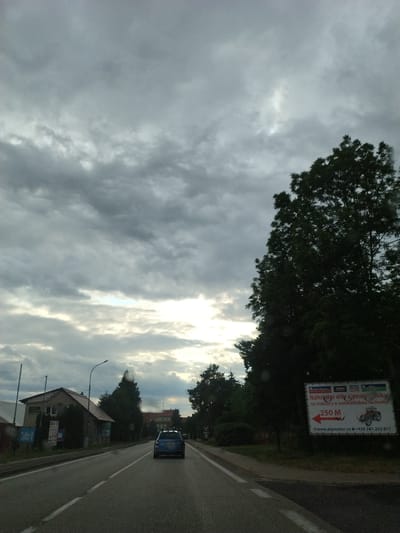 The image shows a cloudy sky over a road in Dražice, Czechia. A blue car is driving on the road, and there are several buildings and trees on the side of the road. There is also a billboard with information about spare parts for tractors and agricultural machinery. The billboard also displays a website address and a phone number. The overall atmosphere is somewhat overcast and muted due to the cloudy sky.