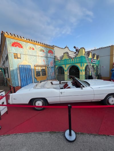This image depicts an outdoor scene featuring a classic white car on a red carpet, set against two vibrantly painted buildings with strong Cuban themes, likely in Fuengirola, Spain.
The central subject is a pristine white classic convertible, identifiable as a Cadillac Eldorado by the badging, presented on a bright red carpet. The carpet is partially cordoned off by a black stanchion with a white base, suggesting an exhibition or display. The car features whitewall tires and chrome hubcaps.
In the background, two buildings showcase elaborate, colorful architecture. The structure on the left has a blue and light blue facade decorated with painted fan-like patterns in red and green. A prominent sign reads "CUBA," and below it, text partially visible includes "Telf. 625 859 421." The building further back on the right features a striking yellow and teal/turquoise color scheme with arched, iron-grilled windows and a painted depiction of the Cuban flag on its upper section. A ladder leans against this building, and a person in a white shirt is visible on its upper level, seemingly working or observing. Another small sign on this building displays "25."
The setting is outdoors on a clear, sunny day, indicated by the bright blue sky with scattered clouds. The overall scene suggests a cultural event, a themed establishment, or a special exhibition celebrating Cuban heritage, using the classic car as a focal point.