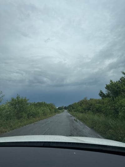 This daytime image, captured in Svoboda, Bulgaria, presents a rural road scene from the perspective of a moving vehicle. A paved, two-lane road, appearing somewhat worn, extends straight into the distance, disappearing at a faint horizon line. Dense green foliage, comprising trees and bushes, lines both sides of the road, creating a natural corridor.
The sky occupies a significant portion of the frame and displays a striking contrast. On the left side, the sky is heavily overcast with voluminous, light gray clouds, indicating cloudy weather. This cloudy expanse abruptly meets a much clearer, darker blue sky on the right side, suggesting a distinct weather front or a rapid change in atmospheric conditions, such as a storm clearing or approaching.
In the immediate foreground, particularly on the right side, a portion of the vehicle's interior frame—likely the dashboard or a window pillar—is visible, confirming the vantage point from inside a car. No people are present in the image, and there is no visible text. The overall scene evokes a sense of travel through a quiet, natural landscape, with the dynamic sky being a prominent and notable detail.