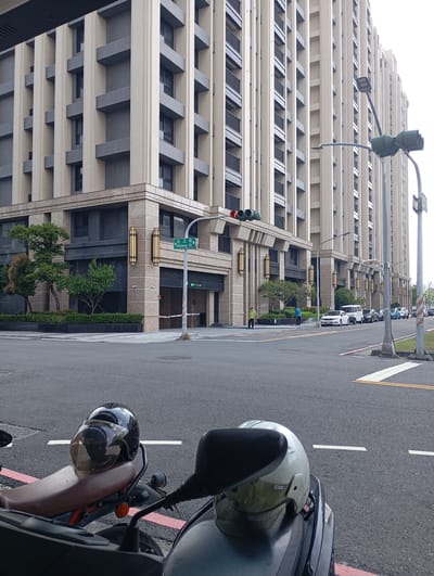 The image captures a street scene in Kaohsiung, Taiwan. In the foreground, two motorcycles with helmets are parked on the street. Behind them, a street intersection leads to a large, modern building with beige and grey architectural details. The building occupies a significant portion of the background. Street signs indicate "Hejiang St". A traffic light is visible at the intersection, showing a red light. Several parked cars and some individuals are present further down the street. The scene appears to be taken during daylight hours. The overall environment is urban, with a focus on the architecture and transportation elements.