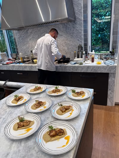 A forensic examination of the image reveals a contemporary kitchen environment, likely a private residence in Naples, United States of America, where a chef is preparing multiple plated dishes.
In the foreground, a light-colored marble kitchen island displays eight white ceramic plates, each featuring an identical meal. The dish consists of a beige puree base, topped with two seared scallops, garnished with microgreens and a thin, dark brown element, and accented by a curved drizzle of orange-yellow sauce on the plate. A black cast iron skillet is visible on the left side of this island.
In the background, a male chef, seen from the back, wears a white chef's jacket and dark pants, with black gloves on his hands. He is actively cooking at a stovetop, stirring contents in a black pan under a large stainless steel range hood. The kitchen backsplash and countertops are fashioned from the same light-colored marble as the island. Various cooking ingredients and utensils, including bottles, jars, bowls of prepped food, and a knife block, are arranged on the counter. Dark cabinetry is installed below the countertop. Large windows to the right reveal dense green foliage, indicating an outdoor view. The overall scene suggests a professional culinary service within a high-end domestic setting.
