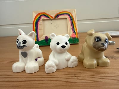 Three plastic animal figurines, a white cat, a white polar bear, and a tan pug, are positioned on a wooden surface. Behind them, a wooden picture frame is decorated with a colorful rainbow and green landscape. The cat has a grey heart on its chest and a smudge of purple on its side. The pug has a dark brown muzzle. The background shows a white wall and possibly a door. The scene is set indoors, on a wooden table, in what appears to be a home environment. There are no people visible in the image. The location in the city of Ridabu, Norway is not discernible from the image.