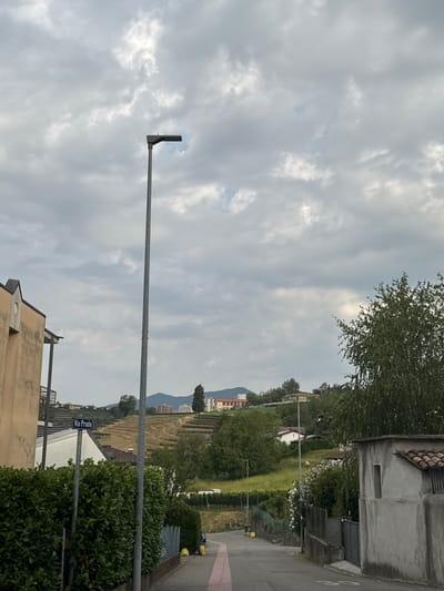 The image depicts a street scene in Balerna, Switzerland, under a cloudy sky. A tall, grey street lamp stands prominently in the foreground, positioned on the left side of a paved road. To the left of the lamp, a portion of a building with a beige-colored facade and dark eaves is visible. A dense green hedge runs along the left side of the road, partially obscuring a metal fence. A blue street sign with white lettering reading "Via Prada" is attached to one of the lamp posts.

The road curves slightly and recedes into the distance, bordered by more hedges and fences. On the right side of the road, there is a grey building with a tiled roof and a small, dark-colored window. Further into the scene, a hilly landscape is visible, characterized by terraced slopes covered in grass and vegetation, interspersed with trees and buildings. The sky is filled with a mix of light and dark grey clouds, suggesting overcast weather. The overall scene is one of a quiet, residential street in a semi-rural setting.