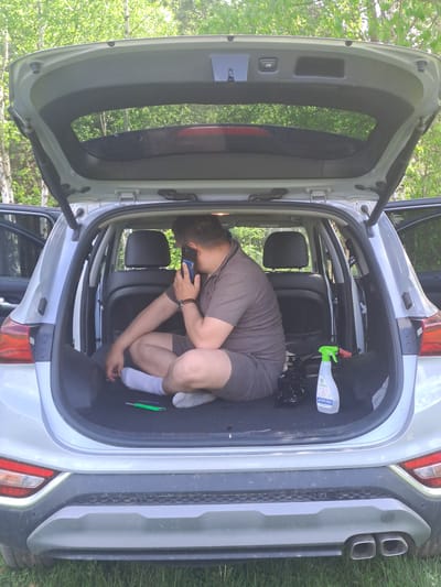 A man is crouched inside the open trunk of a silver car, holding a mobile phone to his ear. He is wearing a short-sleeved brown shirt, brown shorts, and white socks. A bottle with a green spray nozzle is visible near the top of the trunk, along with a black plastic bag. The car is parked outdoors, with green foliage visible in the background. No information suggests the exact location or that it is in Avdot’ino, Russia.