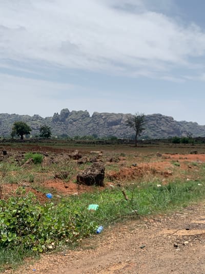 The image depicts a landscape in Makera, Nigeria. A rocky, hilly terrain dominates the horizon. The foreground consists of a dirt road or path, with sparse vegetation on either side. Evidence of human presence is indicated by scattered plastic bottles and debris. The sky is overcast. No individuals are visible in the scene.