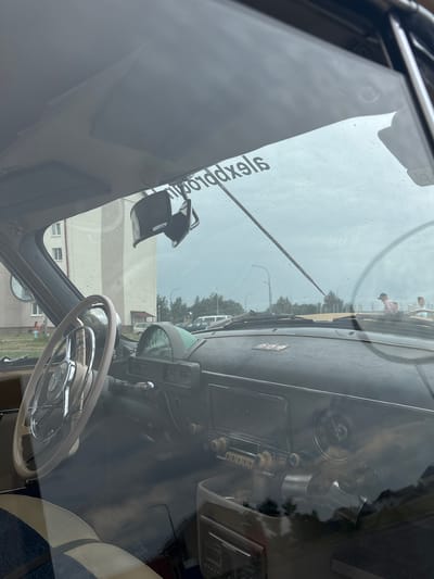 An interior shot of a vintage car, likely a GAZ-21 Volga, is captured through a rain-streaked windshield. The dashboard features a prominent beige steering wheel with chrome accents and a central emblem. To the right, a cluster of vintage gauges includes a circular speedometer and a smaller dial, likely for fuel or oil. Below these, an old-fashioned radio with several knobs and buttons is visible. A hanging rearview mirror is positioned at the top of the windshield, partially obscuring a sticker with Russian text, which reads "pro ddxeig." The exterior scene visible through the windshield includes a modern apartment building with multiple windows, trees, streetlights, and other vehicles, suggesting an urban setting. Two individuals are faintly visible in the distance, one near the apartment building and another near a vehicle on the right side of the frame. The overall atmosphere is overcast and rainy. The scene is set in the city of Uzda, Belarus.