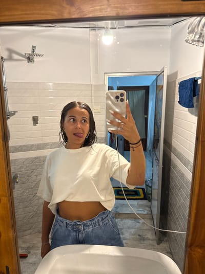 The scene depicts a female individual in a bathroom, situated in Nilwella, Sri Lanka, taking a mirror selfie. The woman has wet, dark hair and is wearing a loose, off-white cropped t-shirt and light blue denim shorts, revealing her midriff. Her right hand holds a light-colored smartphone, equipped with visible camera lenses, pointed towards the mirror. A ring is present on her right ring finger, and her fingernails are painted white. Multiple dark bracelets are visible on her left wrist. She is looking upwards and to her right, with her tongue slightly extended.
The bathroom features walls tiled with horizontal stripes of light grey and white. A chrome shower head is mounted on the wall to the left. A white ceramic sink basin is partially visible at the bottom of the frame. An exposed light bulb in the ceiling provides overhead illumination. To the right, an open doorway reveals an adjacent room with a multi-colored rug on the floor. A blue textured towel hangs on a chrome rack to the right of the mirror. A white electrical cable extends downwards from near the smartphone.