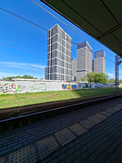 The image depicts a modern urban landscape in Novoivanovskoe, Russia, featuring a row of tall, white apartment buildings with dark window frames against a clear blue sky. In the foreground, train tracks run parallel to a concrete wall covered in vibrant graffiti. The platform, paved with dark bricks and textured tactile paving, is visible on the right side of the frame, partially obscured by an overhang. Lush green grass grows along the base of the graffiti wall. Overhead, electrical wires are strung across the scene, hinting at the presence of a railway station. The overall impression is one of urban development contrasted with artistic expression.