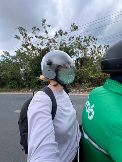 An eye-level outdoor photograph depicts two individuals on a motorcycle riding along a paved road. The person in focus wears a white long-sleeved shirt, a black backpack, a grey motorcycle helmet with a tinted visor, and a light green face mask with white floral patterns. The second person, partially visible behind the first, is wearing a green jacket with the Grab logo. The background includes green trees and bushes, a cloudy sky with power lines, and the asphalt road. Based on the location provided, the scene takes place in Dien Tho, Vietnam.