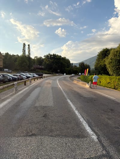 The image depicts a roadside scene in Novalaise, France, with a person standing by the road, looking away from the camera. The person is wearing a light blue t-shirt and red shorts. A wooden railing separates the road from a grassy area. Several cars are parked on the side of the road, and two large yellow banners with red and black text are visible behind the person. The sky is partly cloudy with a bright blue hue, suggesting it's daytime, likely late afternoon or early evening due to the soft lighting. Lush green trees line the background, and a mountain can be seen in the distance. The road itself is asphalt with a white line marking one lane. The overall atmosphere appears peaceful and serene.