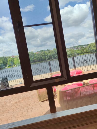 This image captures a scenic view from an indoor space, likely a restaurant or cafe, looking out through a large, angled window. The scene depicts a waterfront area with a body of water, possibly a river or lake, in the mid-ground. A black wrought-iron fence with decorative scrollwork separates the outdoor seating area from the water.
The outdoor area features a paved patio made of terracotta-colored bricks. Several outdoor seating arrangements are visible, consisting of light-colored, woven wicker furniture with bright pink cushions, many of which are still covered in protective plastic. Stacked chairs are also visible near the left side of the patio.
Beyond the fence and water, the landscape is characterized by lush green trees lining the opposite bank. In the distance, a gently sloping hill is dotted with houses and features two tall transmission towers. The sky above is partly cloudy, with large, puffy white clouds against a blue background. The overall impression is that of a pleasant, possibly warm day at a recreational or dining establishment situated by the water.
The location is identified as being in Voskresensky District, Russia, suggesting a connection to the country's natural landscapes and potentially its dacha culture or rural leisure activities.