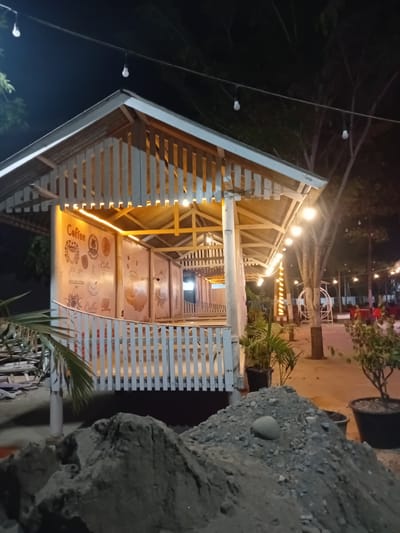The image depicts a nighttime scene at an outdoor cafe in Cotkumbang, Indonesia. The central focus is a light-colored, open-sided gazebo with a sloped roof, illuminated by warm-toned string lights. The walls inside the gazebo are decorated with various coffee-themed graphics. A white picket fence runs along the front of the structure. In the foreground, a large pile of construction debris, likely sand and gravel, is visible. Behind the gazebo, trees and other cafe seating areas with red chairs are present, also dimly lit by string lights. The overall atmosphere suggests a casual, possibly newly established, dining or gathering spot. There are no people visible in the frame.