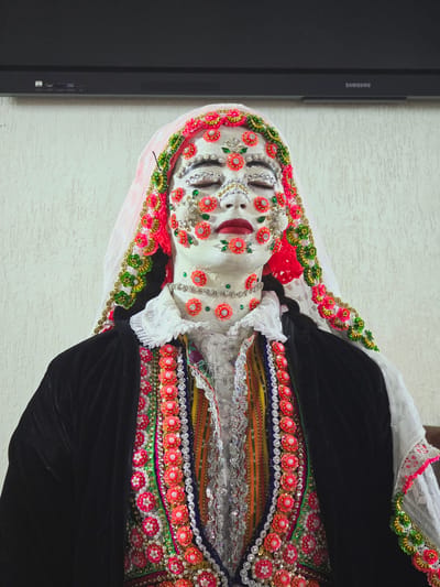 A person wearing elaborate, traditional Ribnovo, Bulgaria attire and makeup is captured in a medium shot. The individual's face is heavily decorated with white makeup and numerous small, colorful floral ornaments and sequins. They wear a dark velvet jacket over a richly embroidered vest adorned with intricate patterns of beads, sequins, and colorful fabrics. A white veil with similar floral embellishments frames their head. The background consists of a textured white wall and a dark electronic device mounted above. The person's eyes are closed, and their expression is serene.