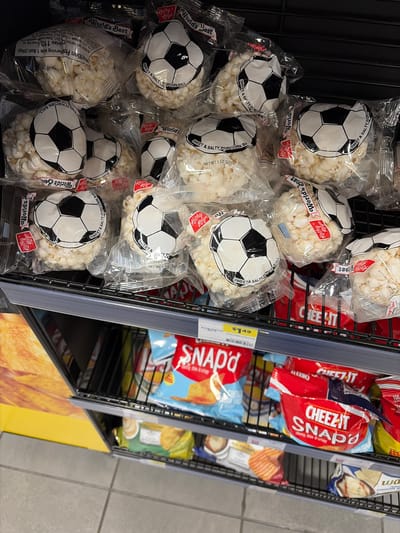 This is a medium shot, eye-level photograph taken from a slightly high angle, showing a display of packaged snacks in a store. The primary subjects are numerous bags of popcorn, each designed to resemble a soccer ball with a black and white hexagonal pattern. Some bags also feature the brand name "World's Best" and "Kathy Kaye."
The popcorn bags are the main focus, filling the left and middle sections of the frame. They are clear plastic bags, allowing the white, puffed popcorn inside to be visible. The soccer ball design is printed on the front of each bag, some appearing slightly faded or creased from being in packaging.
To the right of the popcorn, there is a shelf displaying other snack items. These include red and blue bags of Cheez-It Snap'd snacks, with text such as "cheesy, thin & crispy" visible. Further to the right, partially obscured, are bags of potato chips, one appearing to be Kettle Brand. A yellow price tag with "$1.49" and "Core-Mark Internatio" is attached to the shelf.
The setting appears to be a retail environment, likely a convenience store or grocery store aisle, given the display shelves and the type of products. The lighting is artificial and somewhat flat, consistent with indoor retail lighting. There are no people visible in the image. The overall impression is a commercial display of snack foods.
Visible text includes brand names, product descriptions like "SWEET & SALTY POPCORN BALL," nutritional information snippets, and pricing. The product is sold in the United States, as indicated by the "Troutdale, United States of America" context.