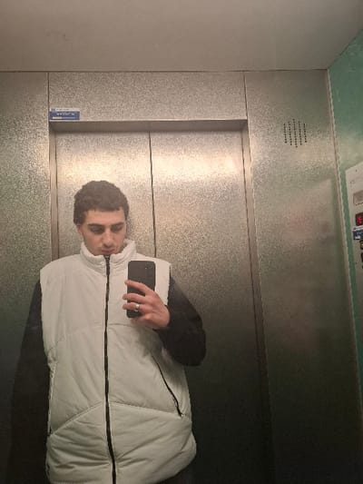 A male individual is depicted in a mirror selfie taken inside an elevator car in Grenoble, France. The elevator walls and doors are constructed from textured, light-colored metallic panels. Above the central elevator door seam, a blue plaque is affixed, partially legible as "GRENOBLE." To the right of the door, a control panel with a red digital display indicates the number "3." The wall section adjacent to the control panel is painted a light green color. The subject, visible from the chest up, possesses short, dark curly hair and is looking down at the smartphone held in his left hand. He wears a white, sleeveless puffer vest over a long-sleeved dark shirt. A silver-toned ring is present on the ring finger of his left hand, which holds a dark-colored smartphone used to capture the image.