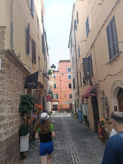 The image depicts a narrow cobblestone street in Alghero, Italy, lined with buildings. On the left, a textured stone wall is visible, adorned with hanging plants in pots. A black flag with white text reading "uno piu uno interiordesign" hangs from a pole. Further down the street, on the right, a woman wearing a white hat, a green shawl, and blue shorts walks away from the camera. She has long, curly brown hair. Across from her, the buildings have facades in shades of beige and pink, with dark shutters on the windows. String lights with bulbs are strung across the street. Several shops are visible, including one with a red awning and a sign that reads "Piadina." A bicycle decorated with colorful flowers is parked on the right side of the street. In the foreground, the back of a man wearing a blue shirt is visible. The overall scene is a typical Mediterranean street with shops and residential buildings.
