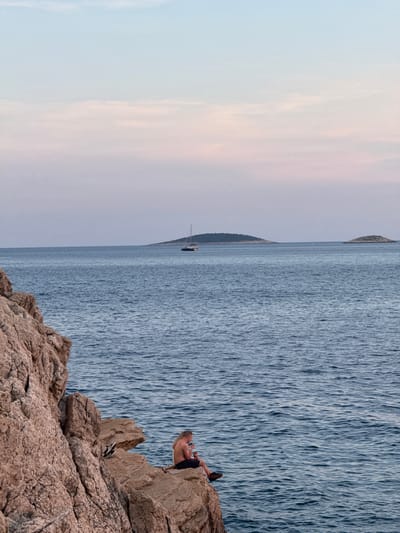 In Primošten, Croatia, a person sits on the edge of a rocky cliff overlooking the Adriatic Sea. The individual, with long blonde hair and bare torso, is wearing dark shorts and appears to be holding a drink. Their legs are crossed, and they are facing towards the right side of the frame, towards the water. To their left, on the rock, are a pair of black shoes. The sea is a deep blue with gentle ripples, and in the distance, a sailboat is visible on the horizon. Two small, tree-covered islands are also present on the horizon. The sky above is a soft gradient of pale blue and pink, suggesting either dawn or dusk. The rocky cliff face, occupying the left side of the image, is textured and reddish-brown in color.
