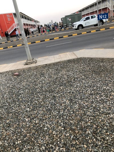 This image depicts an outdoor scene in Hanak, Saudi Arabia, characterized by modular buildings, an asphalt road, and a gathering of individuals.
In the foreground, a large area of mixed-grey and tan gravel covers the ground. A concrete curb transitions to a light grey concrete sidewalk, where the base of a metal light pole is anchored.
The midground features an asphalt road with a painted white line on its surface, bordered by a yellow and black striped curb or barrier. Along the roadside, adjacent to the buildings, approximately 15-20 adult males are gathered. Their attire varies, including traditional Saudi thobes and ghutras, as well as casual clothing. They are observed standing and interacting. A white four-door pickup truck, identified by Arabic script on its side, is parked facing away from the viewer. Behind the truck, a large, olive-green industrial container or generator is visible. A blue sign with white lettering displaying "N12" is positioned near the parked truck. Scattered near the gathering are several bags and small orange cones.
The background primarily consists of modular, multi-story buildings. The closest building, on the left, is red with white exterior staircases and is marked "N12 B5." Further down the road, additional similar modular structures, predominantly light-colored, extend into the distance. The sky above is a light, overcast grey.