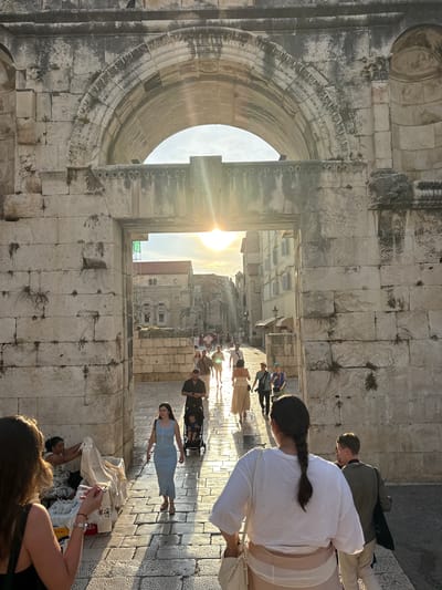 This image captures a sun-drenched scene in Split, Croatia, likely within the ancient Diocletian's Palace. The dominant feature is a massive, weathered stone archway framing a view of a bustling street lined with old buildings. The time of day appears to be late afternoon or early evening, with the low sun casting long shadows and creating a warm, golden light that streams through the archway, creating a lens flare effect.
Several people are visible in the frame. In the foreground, a woman with dark hair in a braid is wearing a white top and a cream-colored skirt, holding a bag. To her right, another woman in a light blue dress walks away from the camera. Further into the scene, several other individuals are walking along the stone-paved street, some appearing to be tourists. One person is pushing a stroller. A vendor is visible at the lower right of the frame, displaying textiles.
The stone architecture is characteristic of Roman or ancient construction, with rough-hewn blocks and decorative carvings around the arch. The street appears paved with cobblestones. The overall mood is relaxed and active, suggesting a pleasant day for exploring historical sites. The bright sunlight and clear sky imply fair weather. No visible text is discernible in the image.