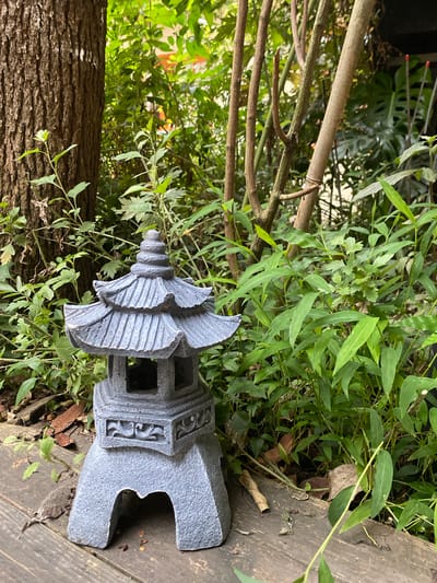 This media file depicts an outdoor scene with a focus on a gray, pagoda-shaped garden ornament. The ornament is made of a rough-textured material, possibly concrete or stone, and features multiple tiers with decorative carvings. It is positioned on a wooden surface, which appears weathered and slightly uneven, suggesting an outdoor deck or walkway.

Surrounding the ornament is lush green foliage, with various types of plants and leaves visible. Some of the plants are low-lying, while others extend upwards, creating a natural, somewhat overgrown garden setting. The background includes a large tree trunk with rough bark, adding to the natural and rustic feel of the environment. Thin, brown branches from other plants or trees crisscross the scene.

The lighting suggests it is daytime, with natural light illuminating the scene, casting soft shadows. The overall mood is peaceful and tranquil, typical of a garden setting. There is no visible text in the image. The location context is Dujiangyan City, China, which aligns with the aesthetic of a traditional Asian garden.
