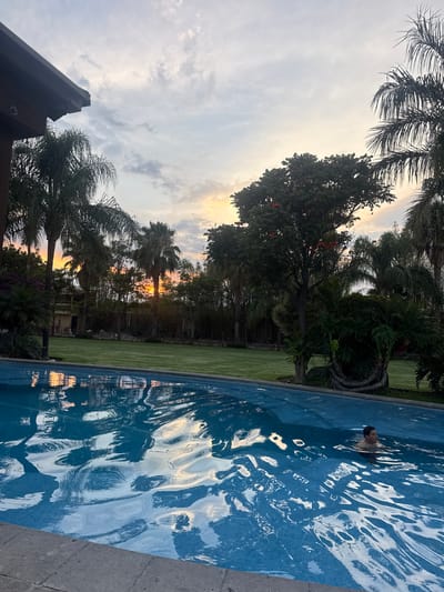 A landscape photograph taken in Santa Ana Yancuitlalpan (Chilhuacán), Mexico. The image shows a swimming pool filled with rippling water, reflecting the sky and trees. A person's head is visible in the water. Trees, including palm trees, surround the pool and a patch of green grass. The sky is visible in the background, with light suggesting it could be near sunrise or sunset. The edge of a building is visible in the bottom left corner.