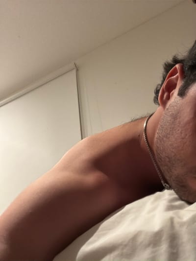 A close-up shot captures a person from an overhead perspective, focusing on their neck, shoulder, and part of their face. A silver chain rests around their neck just above the collarbone. The individual has dark hair and stubble. White bedsheets are visible in the upper right corner. A white wall and a window frame are visible in the lower portion of the image. The lighting is soft and warm.