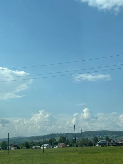 The image depicts a rural landscape in Yugo-Kamskiy, Russia, under a bright, partly cloudy sky. In the foreground, a vast field of green grass stretches out, with some wildflowers visible. Several wooden utility poles with power lines are scattered across the field. In the middle ground, a small village is nestled amongst rolling hills and lush green trees. The village consists of various houses, some with dark roofs and wooden exteriors, and others with lighter colored walls and grey roofs. The background is dominated by a dense forest that covers the undulating hills, giving way to the expansive sky above. The sky is a vibrant blue, with scattered cumulus clouds creating a sense of depth and texture. The overall scene is peaceful and idyllic, characteristic of a summer day in the Russian countryside. There are no people visible in the image.