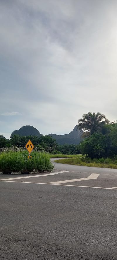 The image depicts a road in Ban Klang, Thailand. The road is paved with asphalt and features white lane markings. A yellow traffic sign indicating a two-way road ahead is visible on the roadside, partially obscured by tall grass. In the background, a mountain range is visible, with a mix of trees and vegetation covering its slopes. The sky is overcast with heavy clouds.