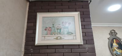 The image shows a framed picture of flowers and decorative objects on a mantelpiece, hanging on a brick wall. To the right of the brick wall, a decorative oval frame with a painting of a woman in historical attire is visible. The environment appears to be indoors, possibly in a home or a museum. No specific location in Ganja City, Azerbaijan can be determined from the image.