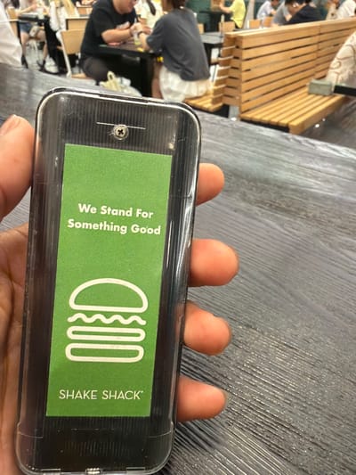 A person's hands are holding a clear phone case with a green insert. The green insert displays the Shake Shack logo, which is a stylized hamburger bun with three wavy lines inside, and text that reads "We Stand For Something Good" and "SHAKE SHACK". In the background, several people are visible in a brightly lit indoor setting, possibly a restaurant or public space. Some people are sitting at wooden benches and tables, and others appear to be standing or walking around. One person is wearing a black t-shirt, and another is wearing a yellow shirt. The scene suggests a casual, everyday environment. The overall impression is a snapshot of a person holding their phone in a public place, with the background showing typical activity in such a location. The image is taken from a close-up perspective on the phone and hands, with the background out of focus.
