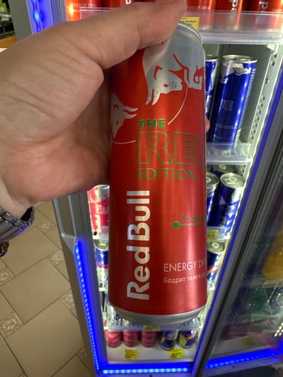 A person's hand holds a can of Red Bull "The Red Edition" energy drink in front of a refrigerated display case stocked with various energy drinks. The can is primarily red with silver lettering and graphics. The background shows shelves of other energy drink cans, including blue cans labeled "BLUE" and pink cans. Blue LED lights illuminate the display case. The floor in the foreground appears to be tiled. The scene suggests a retail environment, likely a convenience store or supermarket, located in Mikhailovka, Russia, as indicated by the context provided.