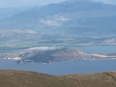 This is a landscape photograph taken from a high vantage point, overlooking a bay and a mountainous region. In the foreground, a dry, grassy hillside with scattered rocks slopes downwards. The middle ground is dominated by a large, dark blue body of water, likely a bay or sea. Jutting into the water is a peninsula covered in scrubby vegetation, with a cluster of white buildings visible on its western tip. A thin band of hazy white clouds hovers over the highest point of this peninsula.

Beyond the peninsula and the water, the landscape rises into rolling hills and distant mountains. These mountains are layered in shades of blue and grey due to atmospheric perspective, with the furthest peaks appearing almost white. Some areas of the hills show signs of cultivation, with green fields visible. Small clusters of buildings are sparsely scattered across the lower slopes of the mountains. The overall impression is one of a serene, natural landscape with some human habitation. The light suggests it is daytime, possibly late morning or early afternoon, with a slightly hazy or overcast sky. There is no visible text in the image.